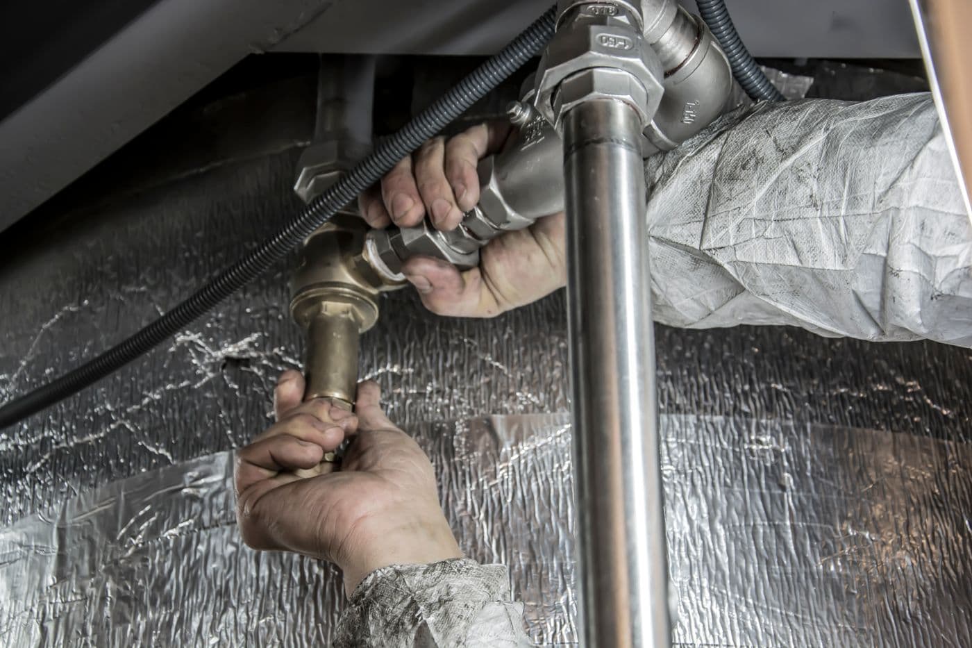 Two bare hands tightening the compression nut on a chrome sink trap in the confined space under a basin, with insulation behind the pipework.